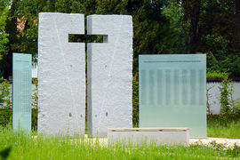 Dreiteilies Mahnmal: Mittig aus Stein mit Aussparung in Form eines Kreuzes, flankiert von zwei Glaswänden mit den eingravierten Namen der Opfer