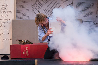 Szene aus einer Aufführung von Kulturmobil. Ein Mann kniet am Boden, in Hand hält er mehrere Blätter Papier. Vor ihm Rauch und Feuer. 